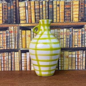 May include: A white ceramic vase with a green checked pattern. The vase has a small handle and a rounded body. The background features a bookshelf filled with books.