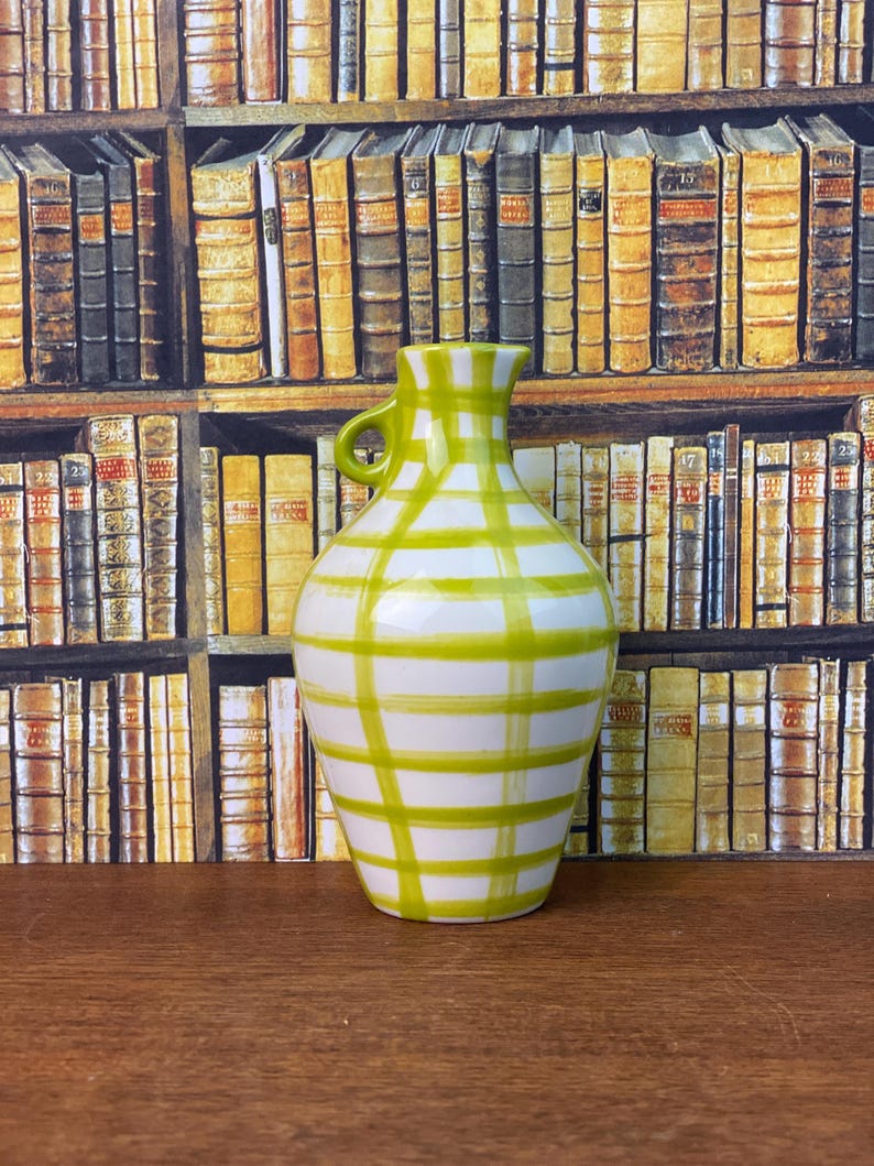 May include: A white ceramic vase with a green checked pattern. The vase has a small handle and a rounded body. The background features a bookshelf filled with books.