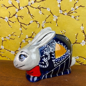 May include: A ceramic rabbit figurine with a silver head and ears, a navy blue body with white and yellow accents, and a red bow tie. The figurine is on a wooden surface with a yellow floral background.
