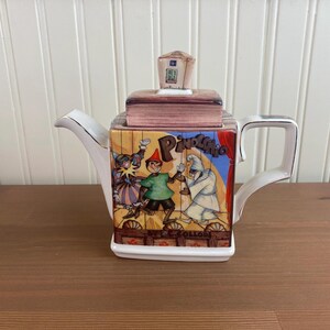 May include: A square ceramic teapot with a Pinocchio theme. The teapot features a colorful illustration of Pinocchio and other characters from the story. The lid is topped with a small house. The handle and spout are white with gold trim.