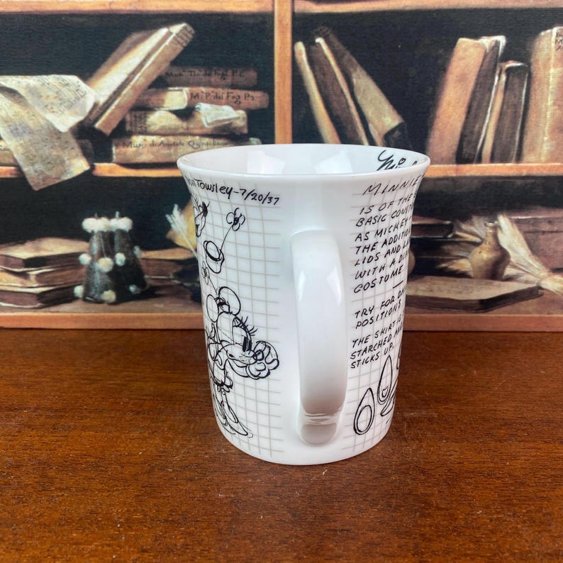 Puede incluir: Taza de cer&aacute;mica blanca con un dise&ntilde;o de boceto de Minnie Mouse en blanco y negro. La taza tiene un asa y texto que dice "Minnie is of the basic..." y "Towsley-7/20/37". La taza est&aacute; sobre una superficie de madera.