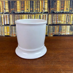 May include: A white ceramic shaving mug with a flared base and a cylindrical body. The mug is set against a backdrop of a bookshelf filled with books. The mug is empty and ready for use.