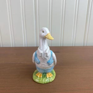 Op de afbeelding: Een keramieken beeldje van een witte eend met een gele snavel en voeten, gekleed in een blauw vest en broek. Het staat op een groene, grasachtige basis. Een decoratief item.