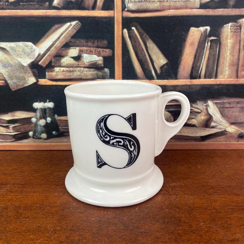 May include: A white ceramic mug with a black, ornate letter "S" on the front. The mug has a small handle and a wide, stable base. The background features a bookshelf with books.