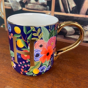 May include: A ceramic mug with a gold handle and rim, featuring a navy blue background adorned with colorful floral patterns and the gold letter "H". The flowers include orange, pink, and blue blooms. The mug is designed for hot beverages.