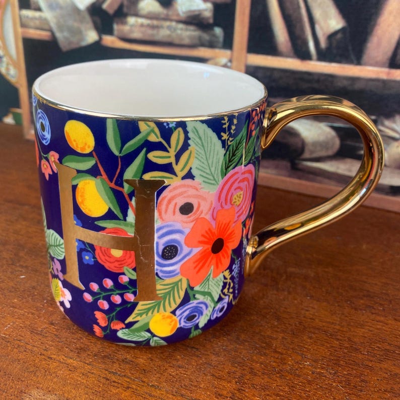 May include: A ceramic mug with a gold handle and rim, featuring a navy blue background adorned with colorful floral patterns and the gold letter "H". The flowers include orange, pink, and blue blooms. The mug is designed for hot beverages.