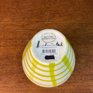 May include: A small, white ceramic bowl with a yellow striped design. The bowl has a rounded shape and a slightly flared rim. The bottom of the bowl has a sticker with the word "ANTHROPOLOGIE" and the text "Made in China Fabriqu&eacute; en Chine".