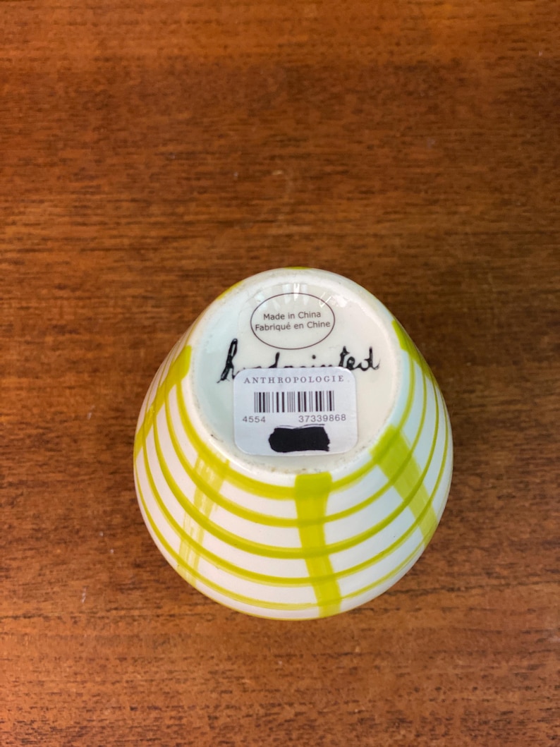 May include: A small, white ceramic bowl with a yellow striped design. The bowl has a rounded shape and a slightly flared rim. The bottom of the bowl has a sticker with the word "ANTHROPOLOGIE" and the text "Made in China Fabriqu&eacute; en Chine".