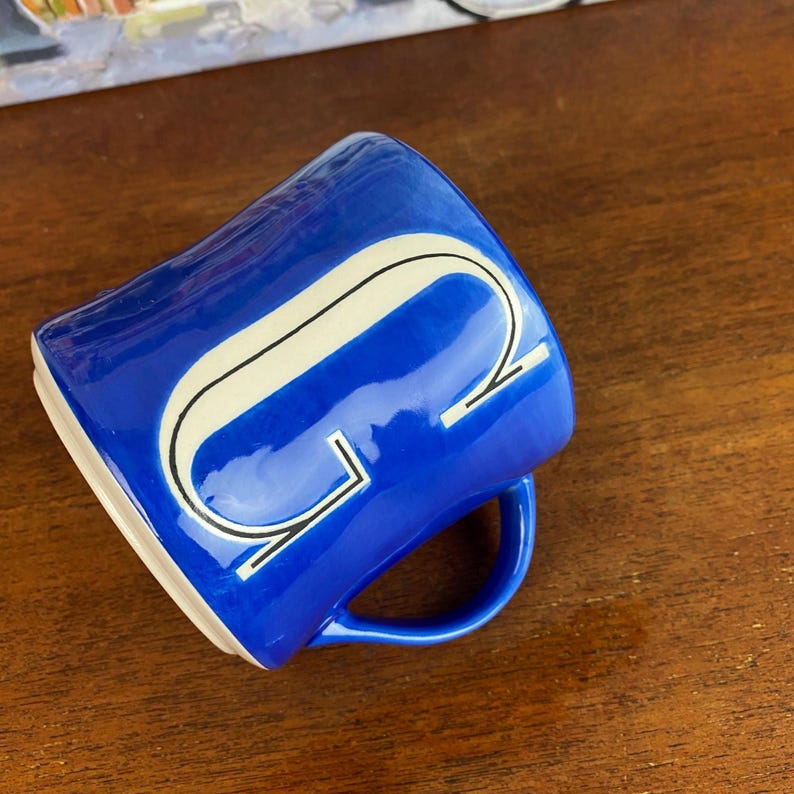 May include: A cobalt blue ceramic mug featuring a white and black stylized letter "G". The mug has a curved handle and a white interior. The mug is placed on a wooden surface.