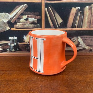 May include: Orange ceramic mug with a white letter "H" on the front. The mug has a curved handle and a white interior. The background features a bookshelf with books.