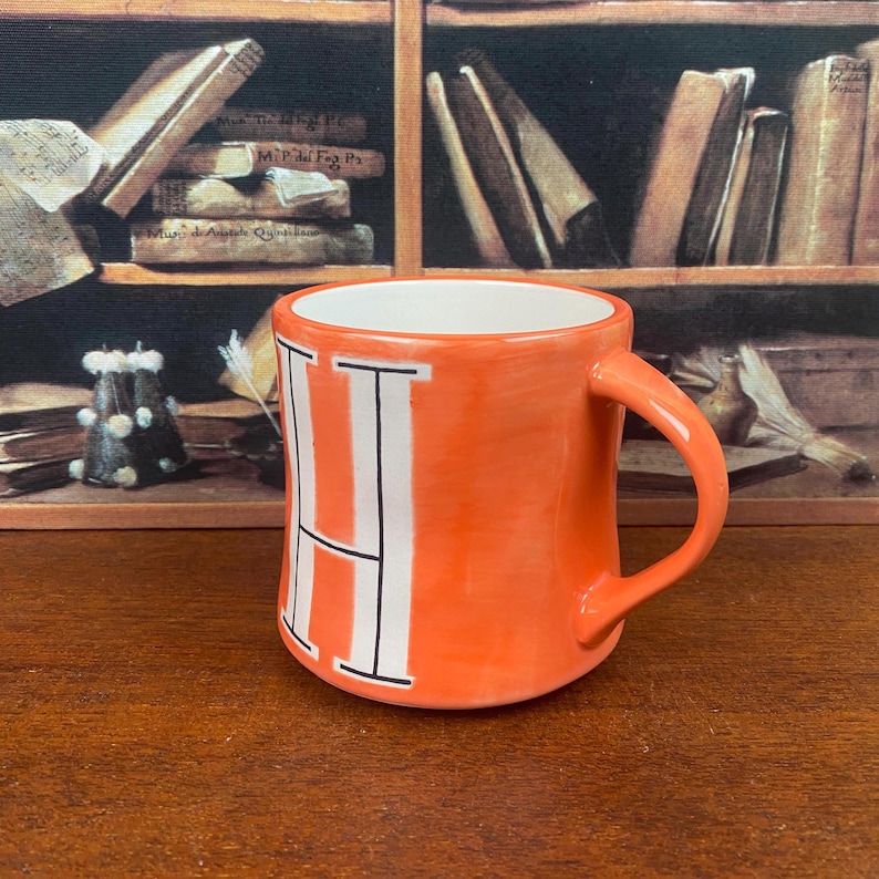 May include: Orange ceramic mug with a white letter "H" on the front. The mug has a curved handle and a white interior. The background features a bookshelf with books.