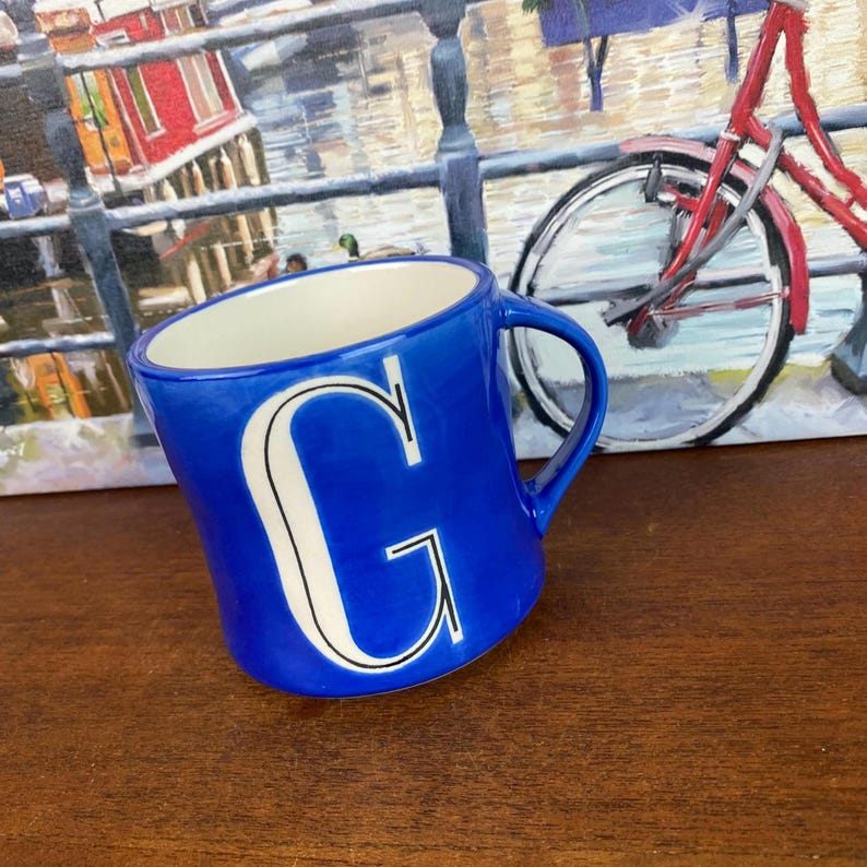 May include: A cobalt blue ceramic mug with a white interior and a large white letter "G". The mug has a curved shape and a handle. The background features a painting of a canal scene and a bicycle.