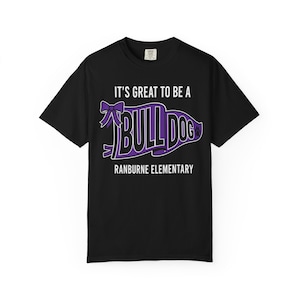 May include: A black short-sleeved t-shirt featuring a purple pennant graphic with a bow. The text reads IT'S GREAT TO BE A BULLDOG and RANBURNE ELEMENTARY in white capital letters. A small paw print icon is visible on the right side of the pennant.