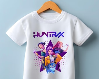 May include: White t-shirt featuring a graphic of three animated figures and a blue cat on a purple star. The word "HUNTR/X" is printed in purple above the graphic. The shirt is hanging on a wooden hanger.