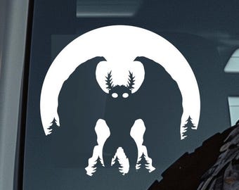 Puede incluir: Una calcomanía de vinilo blanco que presenta una silueta de una criatura parecida a una polilla con grandes alas, sobre una luna creciente blanca. El diseño incluye árboles estilizados en la parte inferior y los lados, creando una imagen espeluznante pero artística.