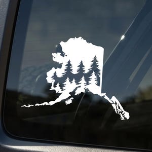 May include: A white vinyl decal applied to a dark vehicle window, featuring the outline of the state of Alaska filled with a silhouette pattern of evergreen pine trees. The design creates a contrast against the dark glass surface.