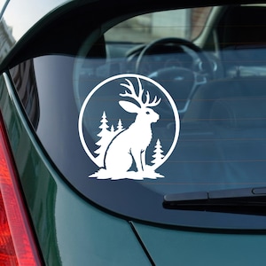 Peut inclure: Un autocollant en vinyle blanc appliqué sur une vitre de voiture représentant la silhouette d'un jackalope, un lapin avec des bois, assis devant un arrière-plan circulaire contenant des sapins. Le motif est rendu en blanc uni sur le verre.