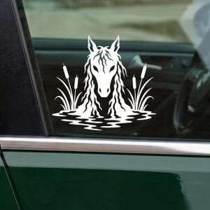 May include: A white vinyl decal featuring the head and flowing mane of a horse emerging from water, flanked by tall cattail plants. The design is rendered in a solid white silhouette style and is applied to a dark car window surface.
