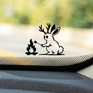 May include: A black vinyl decal sticker featuring a jackalope, a rabbit with antlers, sitting beside a small campfire. The creature is holding a stick with a marshmallow over the flames. The design is a solid black silhouette graphic.