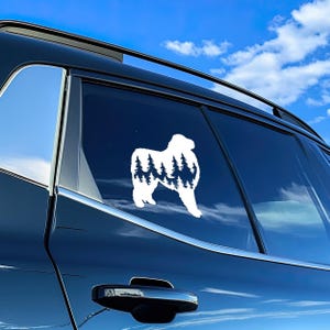 May include: White vinyl decal of a Pyrenean Mountain Dog silhouette with a forest scene inside the dog's body. The decal is on the back window of a dark blue car.
