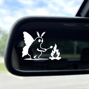 May include: A white vinyl decal applied to a car side mirror, featuring a silhouette of a moth-like creature with large wings and antennae, crouching down to roast a marshmallow on a stick over a small, flickering campfire flame.