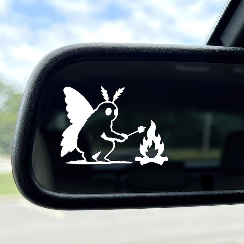 May include: A white vinyl decal applied to a car side mirror, featuring a silhouette of a moth-like creature with large wings and antennae, crouching down to roast a marshmallow on a stick over a small, flickering campfire flame.
