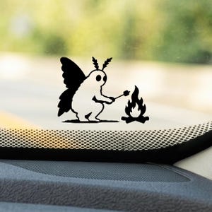 May include: A black and white vinyl decal featuring a moth-like creature with large wings and antennae, sitting and holding a stick over a small campfire. The design is rendered in a solid, high-contrast graphic style on a transparent background.