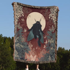 May include: A woven throw blanket featuring a dark moth-like creature with glowing eyes against a full moon. The blanket has a burgundy background with butterflies and foliage in shades of blue, green, and white. The edges are fringed.