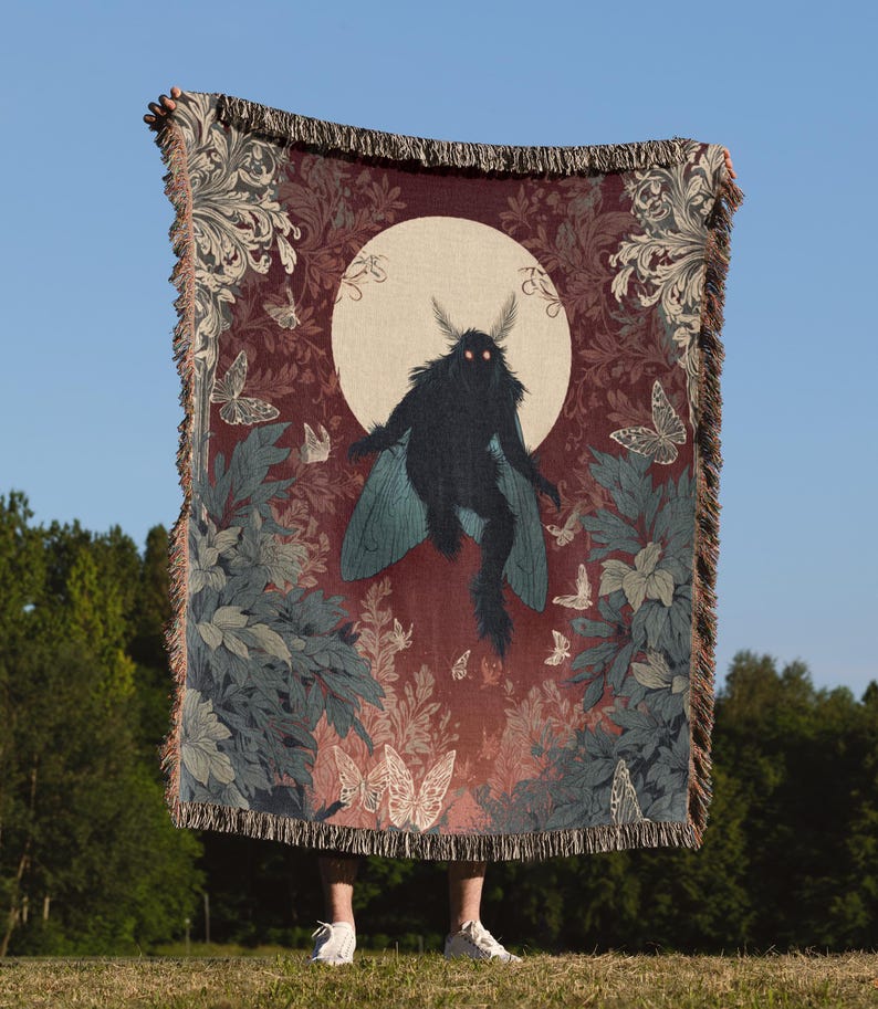 May include: A woven throw blanket featuring a dark moth-like creature with glowing eyes against a full moon. The blanket has a burgundy background with butterflies and foliage in shades of blue, green, and white. The edges are fringed.