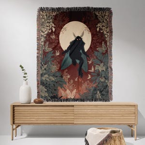 May include: A woven tapestry featuring a dark figure with moth-like wings against a backdrop of a full moon and foliage. The tapestry has a fringed edge and is displayed on a light wood cabinet. Butterflies and floral patterns are also visible.