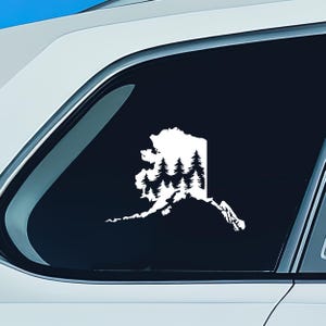 May include: A white vinyl decal applied to a car window, featuring the silhouette of the state of Alaska filled with a detailed graphic of a dense evergreen forest. The design shows multiple rows of pine trees within the state border outline.