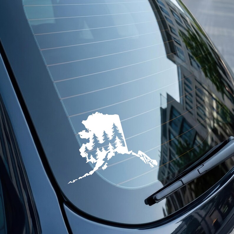May include: A white vinyl decal applied to a car rear window, featuring the silhouette of the state of Alaska filled with a detailed graphic of a dense evergreen forest with multiple pine trees layered within the state borders.