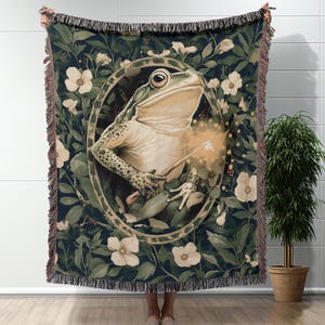May include: A woven tapestry throw blanket featuring a green frog holding a glowing wand in a circular frame surrounded by green leaves and white flowers.