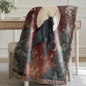 May include: A woven throw blanket featuring a dark red background with a central image of a black moth-like creature against a full moon. The blanket has a fringe and is decorated with butterflies and foliage in shades of green, beige, and brown.