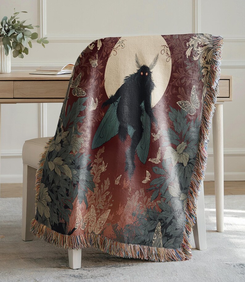May include: A woven throw blanket featuring a dark red background with a central image of a black moth-like creature against a full moon. The blanket has a fringe and is decorated with butterflies and foliage in shades of green, beige, and brown.