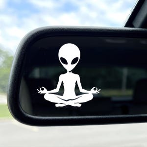 Peut inclure: Un autocollant en vinyle blanc représentant une silhouette extraterrestre stylisée assise en position de méditation, les jambes croisées et les mains en position mudra. Le motif est appliqué sur un rétroviseur extérieur rectangulaire.