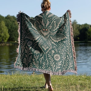 Puede incluir: Una manta verde oscuro con un gran dise&ntilde;o de polilla en el centro, rodeada de patrones florales y bot&aacute;nicos. La manta tiene un borde con flecos con detalles en marr&oacute;n y beige. La persona est&aacute; de pie cerca de un lago.