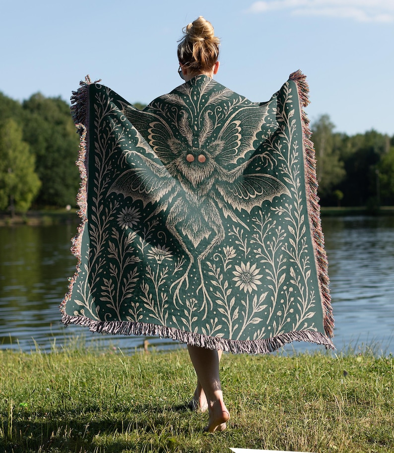 Puede incluir: Una manta verde oscuro con un gran dise&ntilde;o de polilla en el centro, rodeada de patrones florales y bot&aacute;nicos. La manta tiene un borde con flecos con detalles en marr&oacute;n y beige. La persona est&aacute; de pie cerca de un lago.