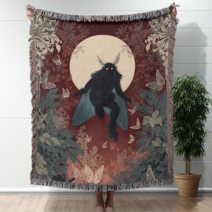 May include: A woven throw blanket featuring a dark moth-like creature with glowing red eyes, set against a backdrop of a full moon and butterflies. The blanket has a burgundy background with floral and foliage patterns, and a fringed edge.