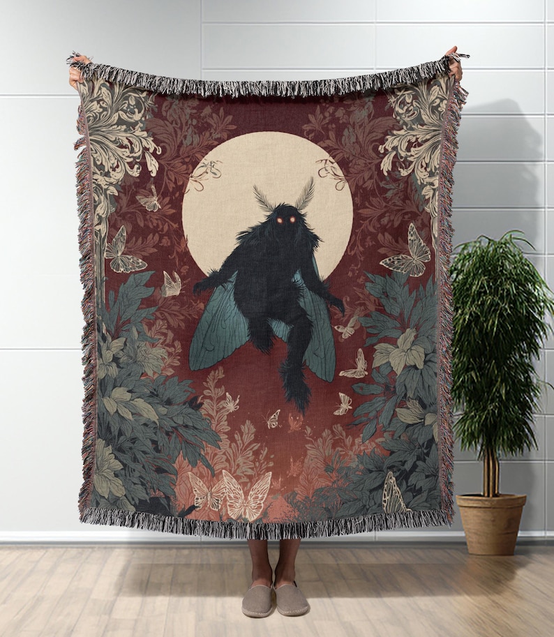 May include: A woven throw blanket featuring a dark moth-like creature with glowing red eyes, set against a backdrop of a full moon and butterflies. The blanket has a burgundy background with floral and foliage patterns, and a fringed edge.