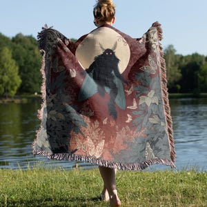 May include: A burgundy throw blanket with a large moth illustration against a full moon. The blanket features a fringe edge and a pattern of butterflies and foliage in shades of gray and green. The person holding the blanket is standing near a lake.