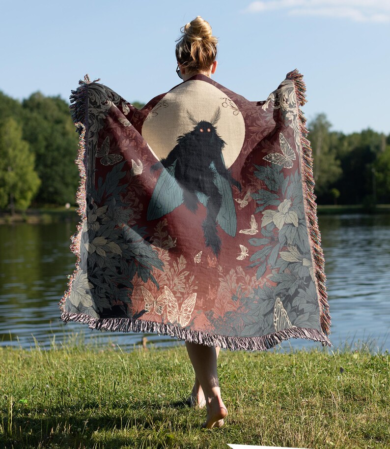 May include: A burgundy throw blanket with a large moth illustration against a full moon. The blanket features a fringe edge and a pattern of butterflies and foliage in shades of gray and green. The person holding the blanket is standing near a lake.