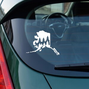 May include: A white vinyl decal applied to a car rear window, featuring the silhouette outline of the state of Alaska filled with a detailed graphic of a dense evergreen forest with multiple pine trees layered within the state borders.