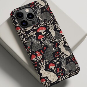 May include: Black phone case with a repeating pattern of white rabbits with antlers, red mushrooms, and green leaves.