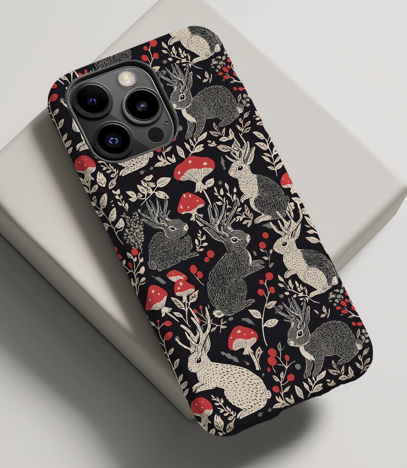 May include: Black phone case with a repeating pattern of white rabbits with antlers, red mushrooms, and green leaves.