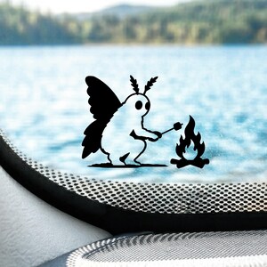 May include: A black vinyl decal featuring a moth-like creature with large wings and antennae, sitting and roasting a marshmallow on a stick over a small campfire. The design is solid black and is applied to a transparent surface.