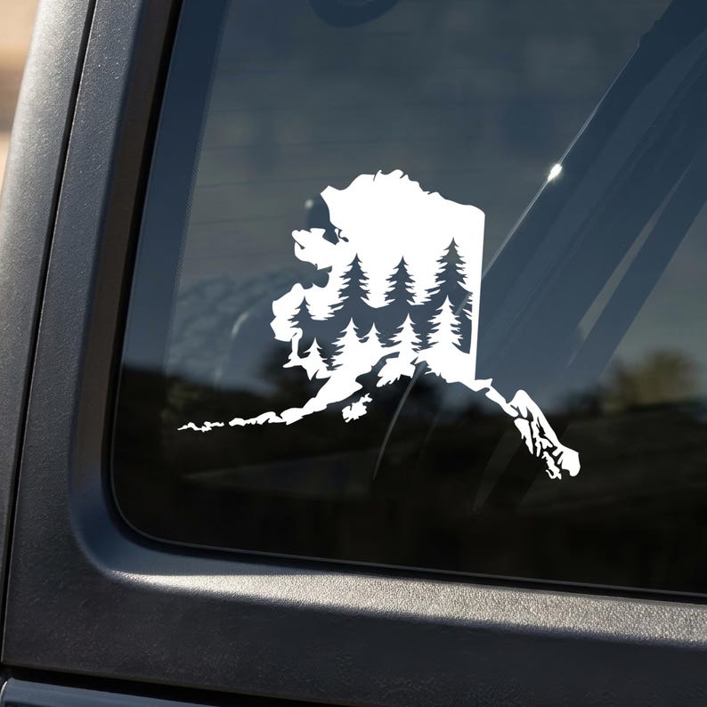 May include: A white vinyl decal applied to a dark vehicle window, featuring the outline of the state of Alaska filled with a silhouette pattern of evergreen pine trees. The design creates a contrast against the dark glass surface.