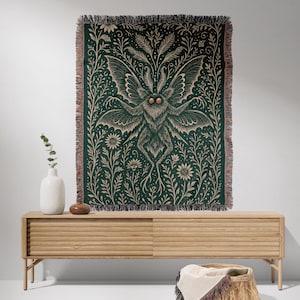 Puede incluir: Manta tejida verde oscuro con una ilustraci&oacute;n detallada de una polilla y detalles florales. La polilla tiene grandes alas y est&aacute; centrada en la manta. La manta tiene un borde con flecos y se exhibe sobre un mueble de madera clara.