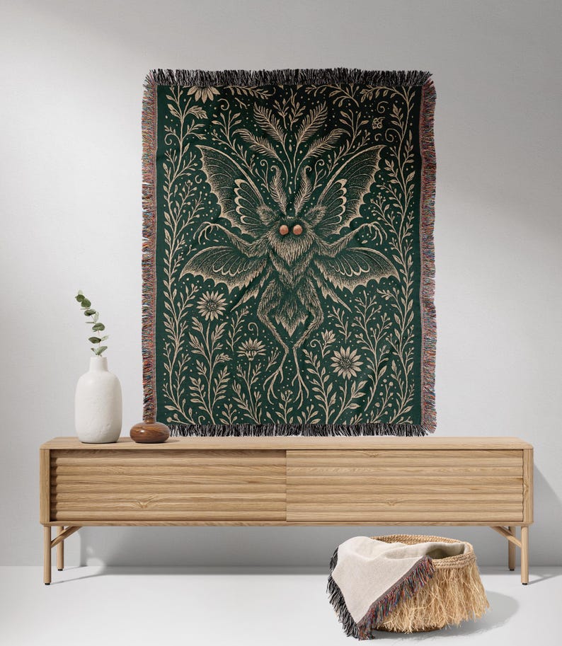 Puede incluir: Manta tejida verde oscuro con una ilustraci&oacute;n detallada de una polilla y detalles florales. La polilla tiene grandes alas y est&aacute; centrada en la manta. La manta tiene un borde con flecos y se exhibe sobre un mueble de madera clara.
