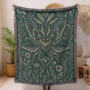 Puede incluir: Una manta tejida de color verde oscuro con un gran dise&ntilde;o de polilla y detalles florales y de hojas en beige claro. La manta tiene un borde con flecos y est&aacute; sostenida por una persona.